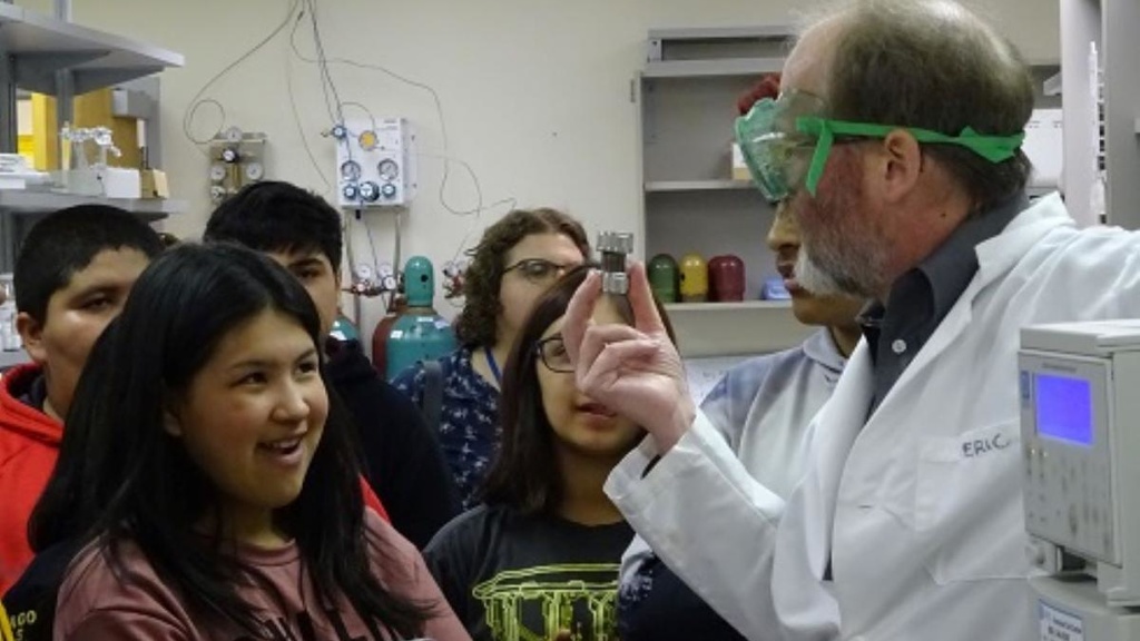 A man in a lab coat and safety goggles talks to a group of youth.